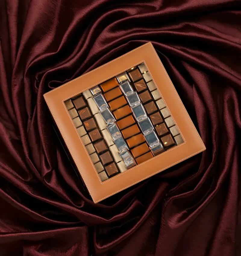 Coffret vitrine en cuir camel rempli de chocolats posé sur un satin bordeaux